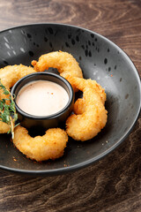 Breaded fried shrimp served on a black plate with dipping sauce and herb garnish, presented on a dark wooden surface. Gourmet appetizer, restaurant-style plating.