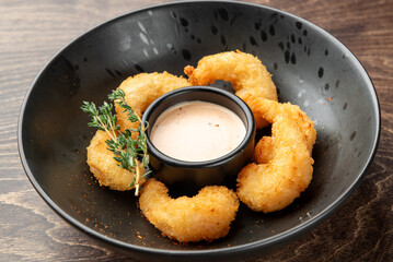 Breaded fried shrimp served on a black plate with dipping sauce and herb garnish, presented on a dark wooden surface. Gourmet appetizer, restaurant-style plating.