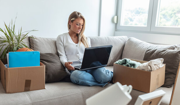 Happy young Caucasian woman using the Moving App on her laptop, surrounded by cardboard boxes and furniture in the background.