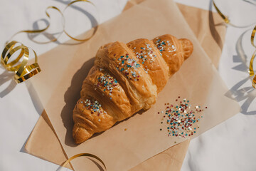 Golden, flaky croissant sprinkled with colorful sprinkles, resting on parchment paper, festive treat.