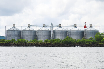 Fototapeta premium Row of large metal grain silos sits on a rocky shore,