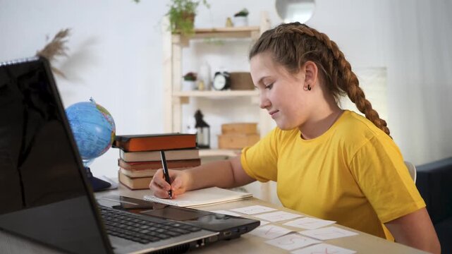 girl studying at desk with laptop and notebook, teenager writing homework note beside globe and stack of book, focused on learning and study, organizing flashcard and note for school assignment