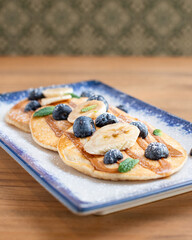 Pancakes with blueberries, banana slices, and a caramel drizzle, dusted with powdered sugar, creating a delicious breakfast plate