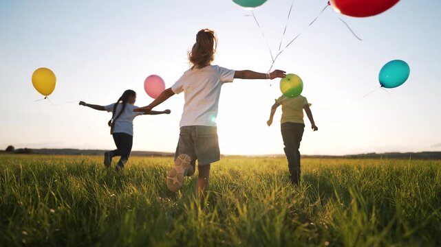 Children run holding colorful balloon across sunlit field and grass at sunset while child play and run in group outdoor summer joy carefree laugh as balloon float over green meadow and play together