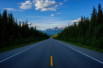 Naklejka premium Scenic Mountain Highway Journey Vast Evergreen Forest Leading Towards Majestic Snow-Capped Peaks Azure Sky White Puffy Clouds Golden Hour Sunlight Dramatic Landscape Adventure Travel Destination