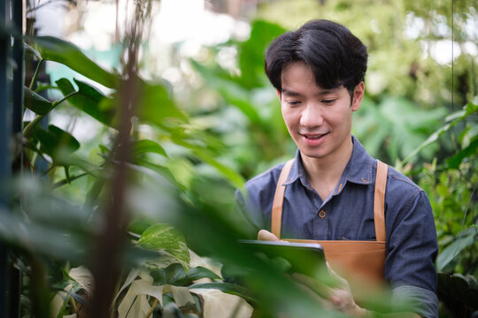 Smiling young man in apron using digital tablet to manage plants in a lush greenhouse setting.
