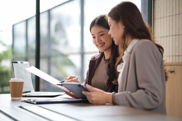Two professional women reviewing paperwork and working on a tablet in a bright modern office.