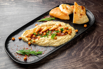 Grilled shrimp served over hummus with chickpeas, tomatoes, herbs, and pita bread on a black plate. Gourmet Mediterranean appetizer, top view, rustic wooden background.