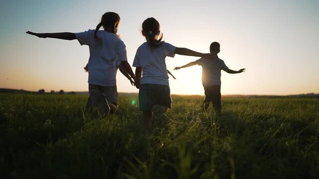 Children run across grassy field at sunset chasing play and laughter under warm sunlight in tall green grass friends holding hand enjoying summer outdoor freedom and friendship in open meadow