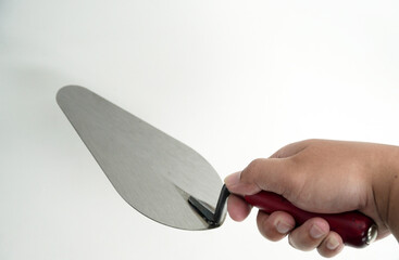 A person's hand firmly grips a professional bricklaying trowel with a red handle, preparing for work. The tool and hand are shown on a clean white background.

