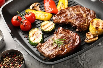 Pan with grilled beef steaks, mushrooms, vegetables and spices on grey textured table, closeup