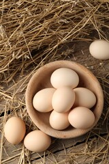 Raw chicken eggs in bowl and straw on wooden table, flat lay