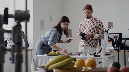 Brunette woman holding tablet while setting table, adjusting plates with dishes. Professional photographer waiting for her while chatting