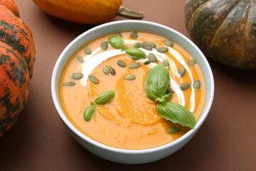 Delicious pumpkin soup with seeds in bowl, basil and vegetables on brown background, closeup
