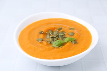 Delicious pumpkin soup with seeds and basil in bowl on white tiled table, closeup