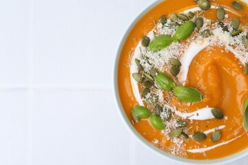Delicious pumpkin soup with spices, cheese and sour cream in bowl on white tiled table, top view. Space for text