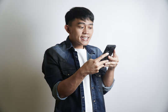 Smiling young Asian man in denim shirt excitedly holding and looking at his smartphone with a surprised and joyful expression, captivated by engaging digital content or an online message