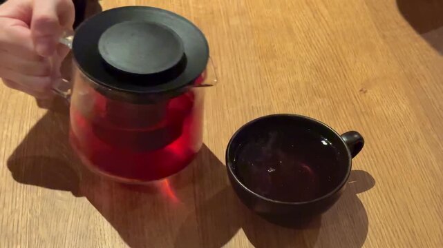 Hot red hibiscus tea is pouring from a glass teapot into black cup, wooden table close-up background