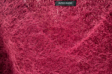 Bright burgundy V neck sweater with a label identifying the English text Alpaca Blend. The surface is fluffy with long fibers, characteristic of alpaca blends.