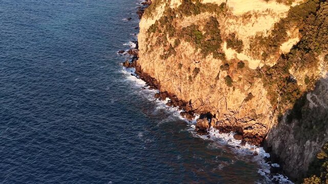 Beautiful cliffs of Punta Imperatore in the sunset with waves crashing on cliffs in coastal island of Ischia in Italy with deep blue sea waves, cliffs and sunny weather. 