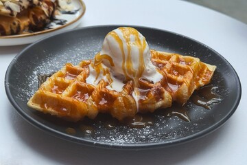 Baked waffle croissant, croffle with vanilla ice cream and caramel sauce topping on a black ceramic plate, close-up food photography