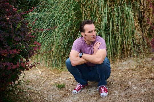 Thoughtful Man Squats Outdoors in Garden Setting With Purple Shirt and Red Sneakers - Powered by Adobe