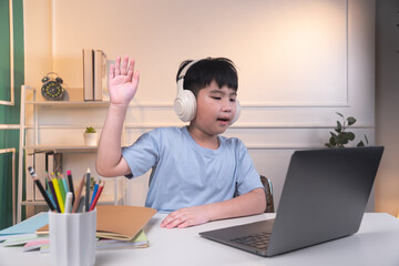Asian boy wearing headphones studying online on laptop is making a gesture of raising his hand.