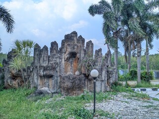 Rocky structure resembling tall pillars with lush green grass, gravel, and tropical palm trees under a cloudy sky.