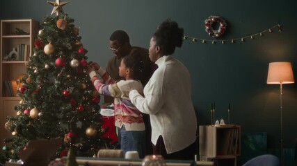 African American family of two adults and girl child decorating artificial fir tree together arranging fairy lights and colorful baubles on branches during Christmas preparation in living room - Powered by Adobe