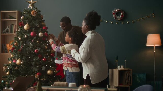 African American family of two adults and girl child decorating artificial fir tree together arranging fairy lights and colorful baubles on branches during Christmas preparation in living room
