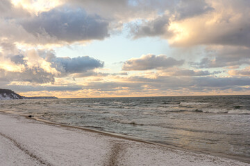 Snow-covered shoreline meets gentle waves under a pastel sky. A tranquil coastal scene with cool tones, soft light, and fluffy clouds evokes a peaceful winter mood, winter beach