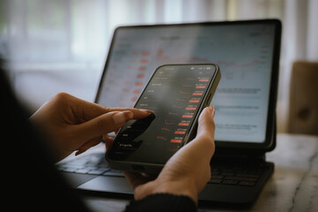 Hands holding smartphone and using laptop with financial graph on screen, showing digital business growth and investment analysis concept.