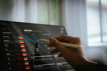 Hand using digital pen on tablet displaying stock market data and falling financial charts,...