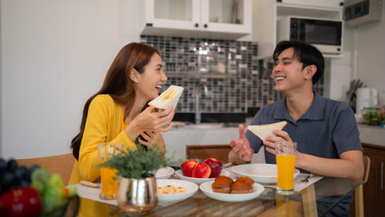 Young Asian adult couple enjoy breakfast eat sandwiches drink juice at home kitchen table sharing happy joyful morning meal bonding connection