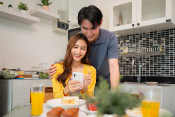 Asian couple share happy morning breakfast home browsing mobile internet social media together kitchen table digital connection fun everyday life