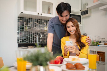 Asian adult couple happy mobile phone moment share affection enjoy leisure breakfast modern home kitchen dining table