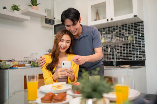 Asian adult couple happy share smartphone home kitchen modern lifestyle digital device connection breakfast joy morning daily routine technology - Powered by Adobe