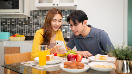 Young Asian couple sharing smartphone browsing internet during morning breakfast at modern kitchen home enjoying digital lifestyle together