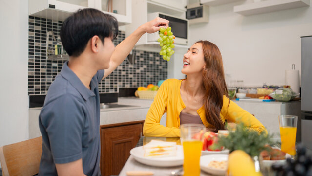 Asian young adult couple enjoys healthy breakfast sharing fresh fruit together home kitchen intimate happy moment domestic scene bright morning family lifestyle - Powered by Adobe