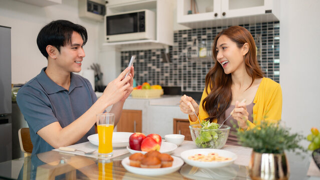 Young Asian couple share joyful meal home kitchen adult man taking photo woman eating salad healthy food happy lifestyle moments