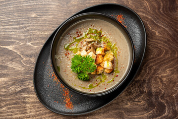Creamy mushroom soup with croutons, parsley, herb oil, and red pepper flakes in a black bowl on a dark wooden table. Gourmet presentation, top view, rustic style.