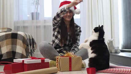 Happy young woman in Santa hat playing with her cat while wrapping Christmas gifts at home. Cozy holiday atmosphere with cute pet, festive mood, and joyful domestic lifestyle. - Powered by Adobe