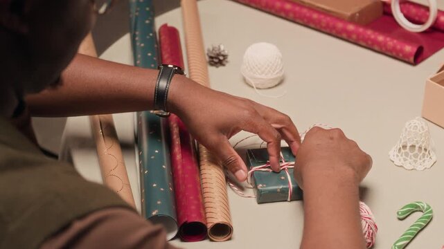 Hands of Black man tying festive twine on green gift box decorating Christmas presents together with family at home while sitting at table cluttered with colorful wrapping papers
