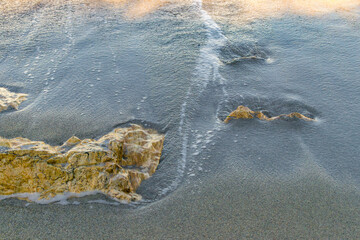 Sea wave washing over yellow rocks on sandy beach at sunset. Close-up of ocean surf texture and...