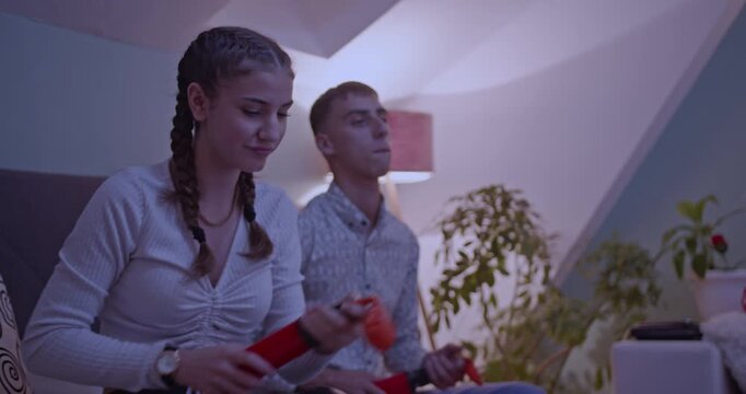 A happy young couple sitting together on a couch, enthusiastically inflating orange balloons with hand pumps. They are laughing and preparing for a cheerful birthday party celebration at home.