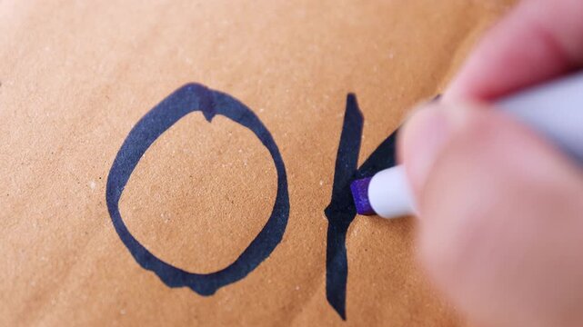 Hand writing "OK" with blue marker on cardboard, symbolizing approval, correctness, or a simple positive message, close-up