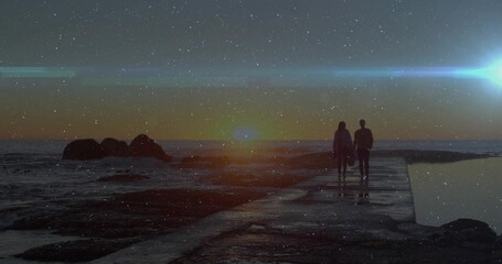 Strolling couple in jackets moving toward glowing sun on concrete pier, with rocks and tidal pool