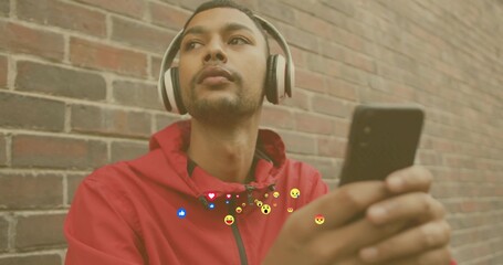Leaning Indian man in red hoodie and headphones checking smartphone on brick wall, floating emojis