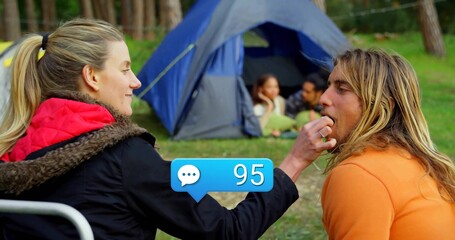Feeding camper wearing red-hooded coat giving snack in pine clearing, with blue tent and green mat