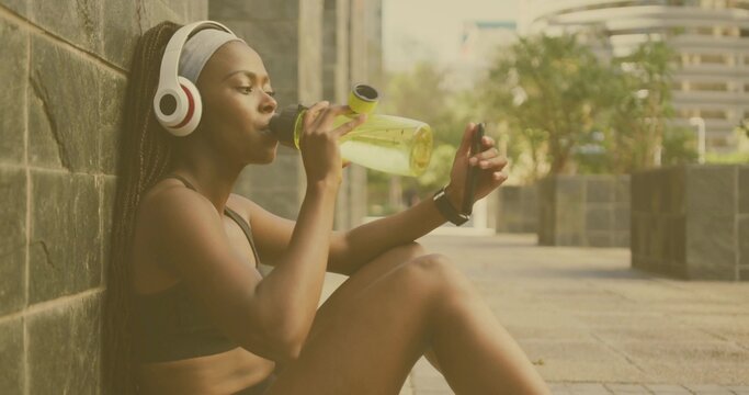 Fototapeta Sitting fitness fan in sportswear drinking from water bottle on plaza, with headphones, copy space